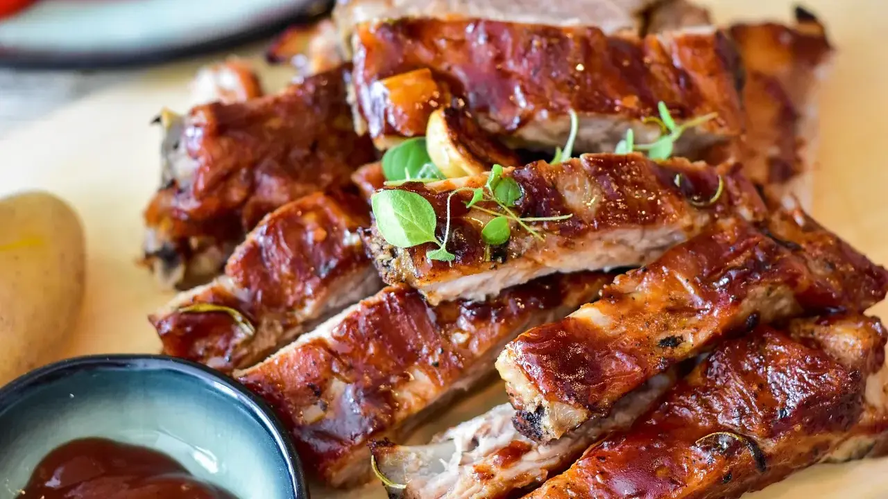 Saftige spareribs backofen, glasiert und mit Kräutern garniert, daneben eine Schale BBQ-Sauce und eine Kartoffel.
