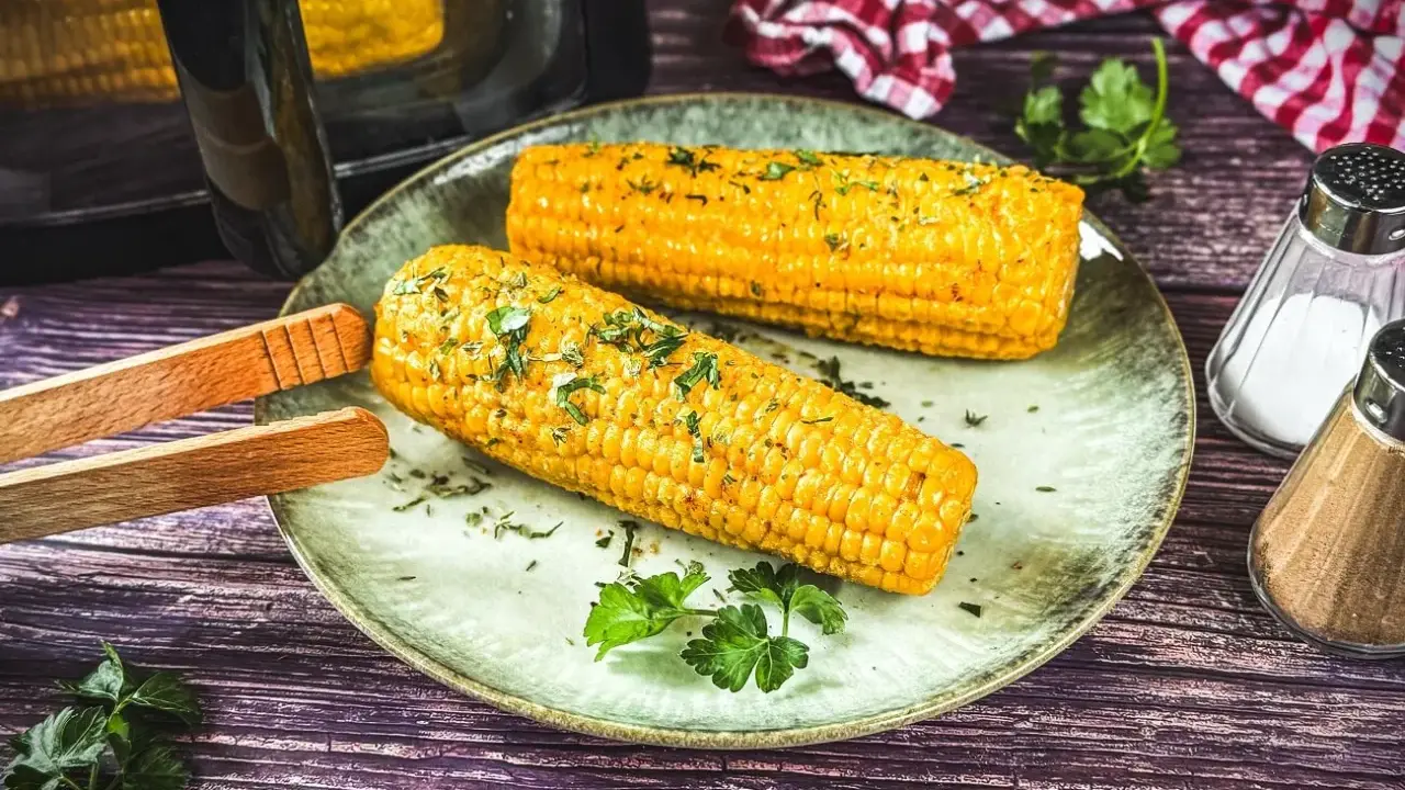Zwei goldgelbe Maiskolben, frisch aus dem Airfryer, liegen auf einem Teller, garniert mit Kräutern. Daneben Salz- und Pfefferstreuer.