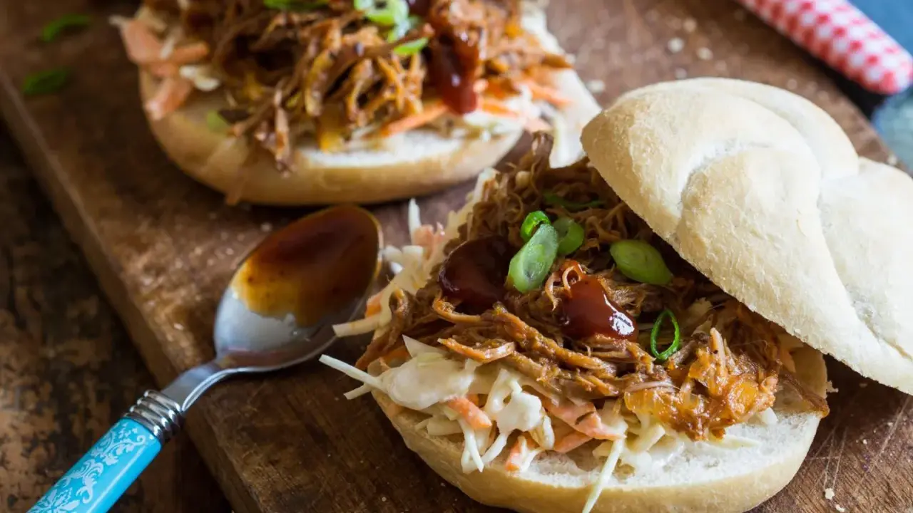 Saftiges pulled pork auf einem Brötchen mit Coleslaw und BBQ-Sauce, garniert mit Frühlingszwiebeln. Ein Löffel mit Sauce liegt daneben.