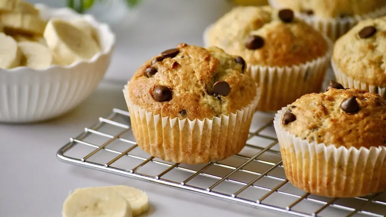 Frisch gebackene schoko bananen muffins mit Schokostückchen auf einem Gitter. Daneben eine Schüssel mit Bananenscheiben.