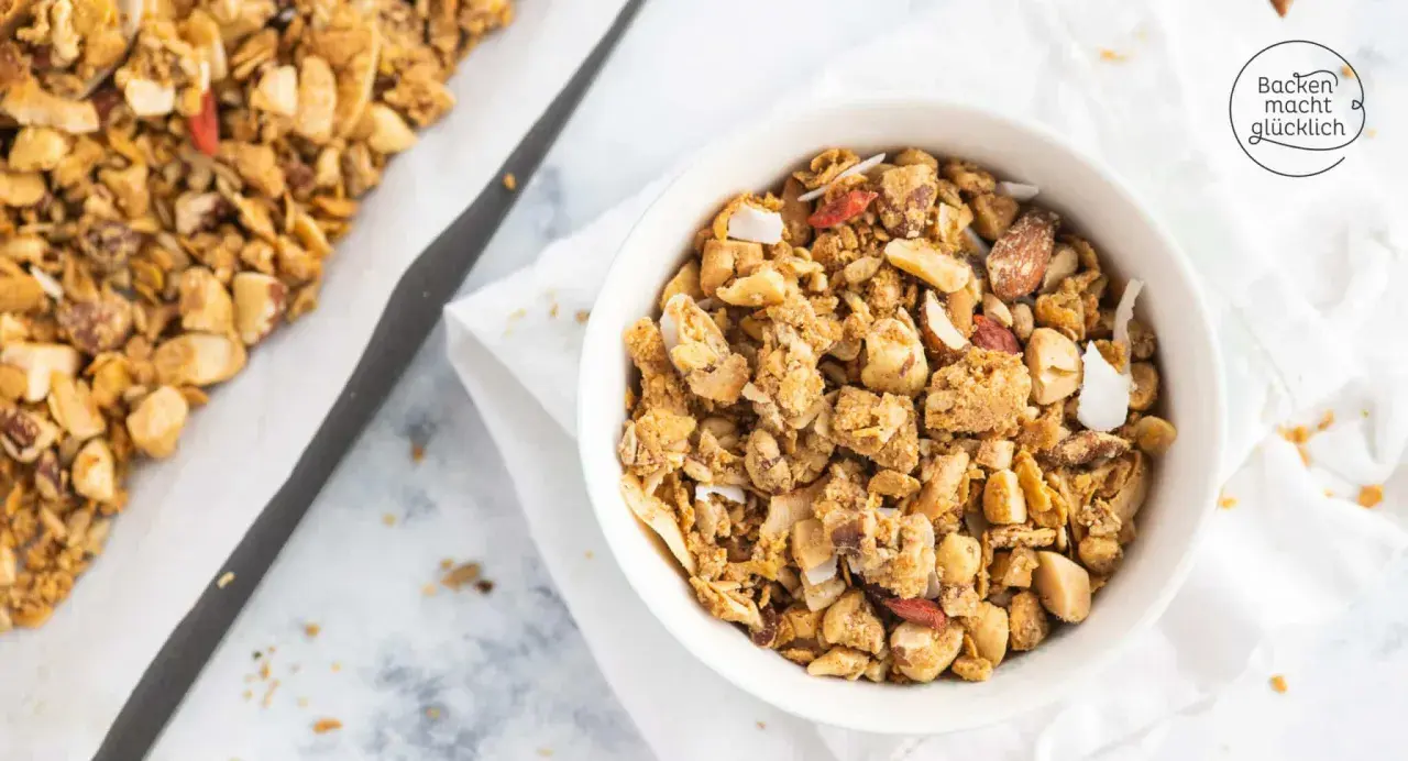 Schüssel mit selbstgemachtem, gesundem Granola, Nüssen und Kokosflocken. Daneben ein Blech mit frisch gebackenem Müsli.