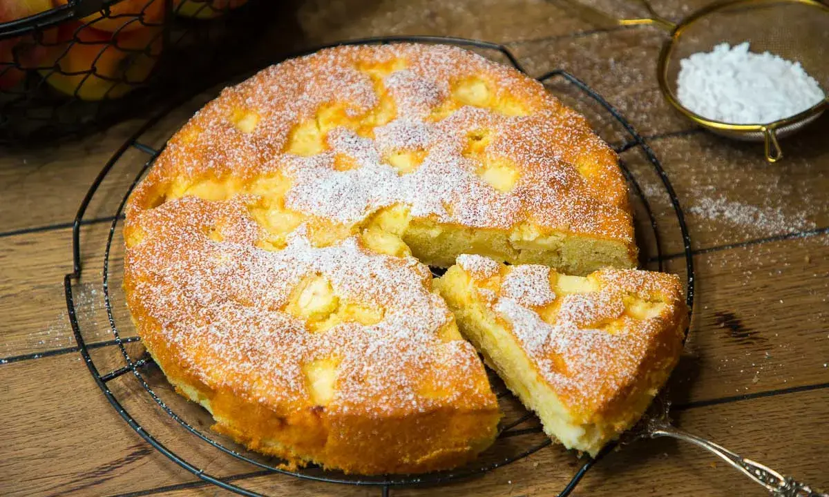 Omas weltbester Apfelkuchen, frisch gebacken und mit Puderzucker bestäubt, wartet auf seine Genießer. Ein Stück ist bereits herausgeschnitten.