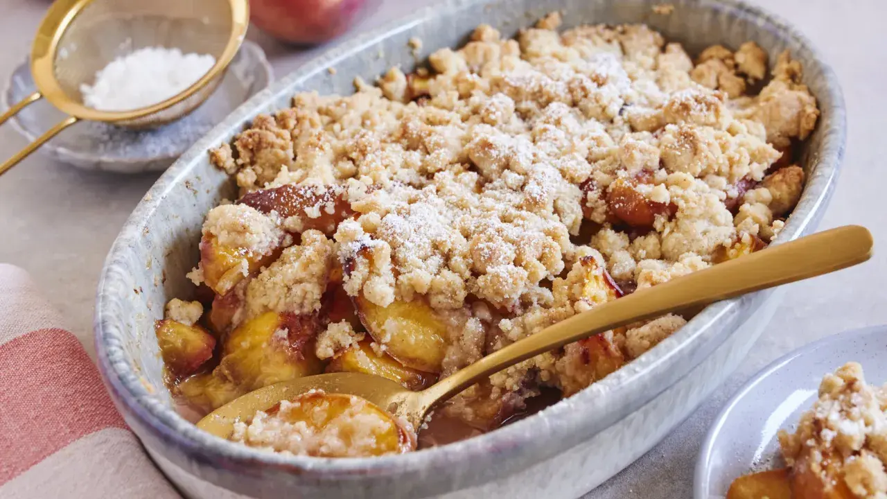 Ein goldbrauner Pfirsich Crumble mit knuspriger Streuseldecke, bestäubt mit Puderzucker. Ein Löffel steckt darin, bereit zum Genießen.