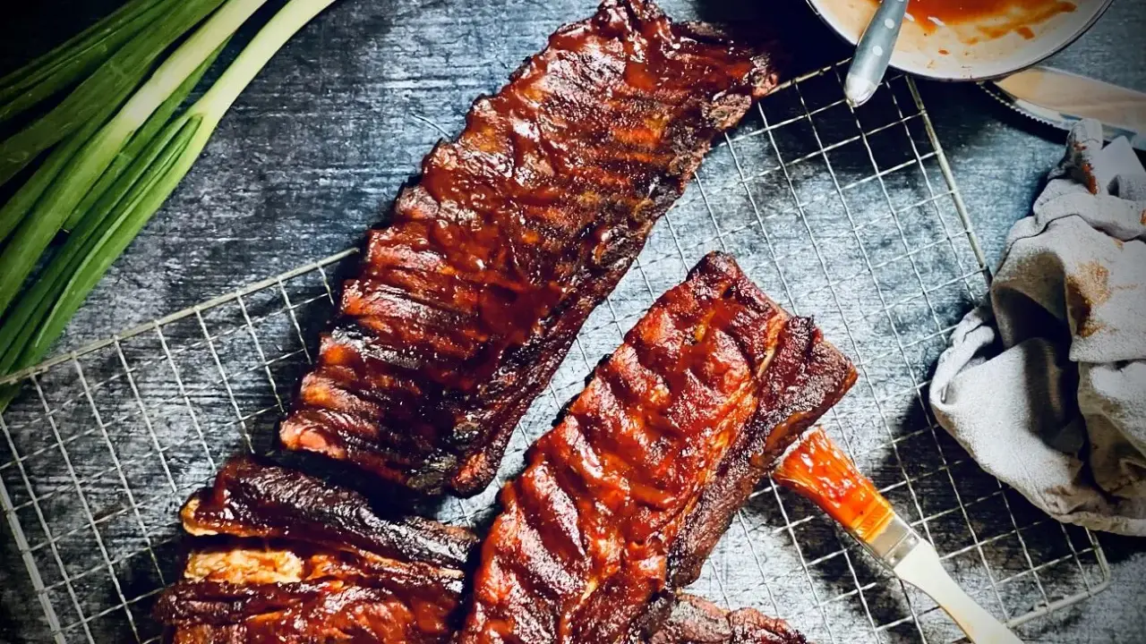 Saftige spare ribs, glasiert mit BBQ-Sauce, auf einem Grillrost. Daneben Frühlingszwiebeln und eine Schüssel Sauce.