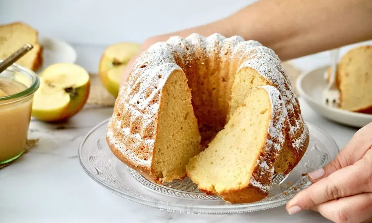 Ein saftiger Kuchen mit Apfelmus, bestäubt mit Puderzucker. Daneben liegen frische Apfelhälften und ein Glas Apfelmus.