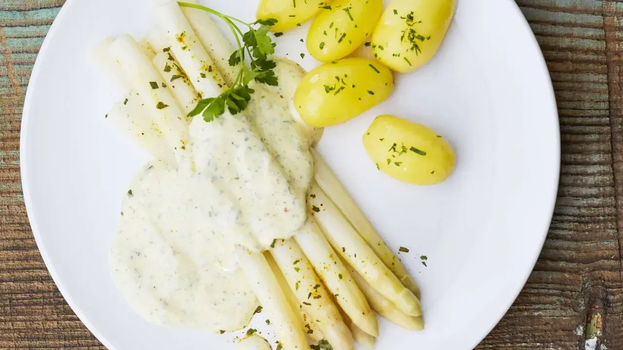 Ein Teller mit weißem Spargel, garniert mit Petersilie und überzogen mit cremiger Hollandaise. Dazu gibt es kleine, goldgelbe Kartoffeln.