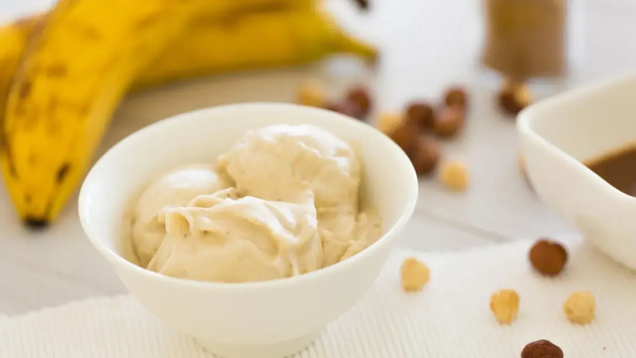 Cremiges Bananeneis, gemacht aus gefrorenen Bananen, in einer Schüssel. Daneben liegen Nüsse und ein Schälchen mit Schokoladensauce.