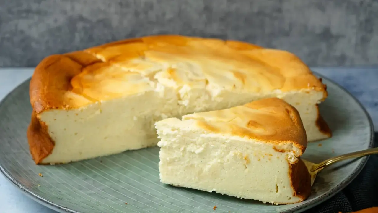 Ein saftiger Quarkkuchen ohne Boden, goldbraun gebacken, liegt auf einem Teller. Ein Stück ist bereits angeschnitten.