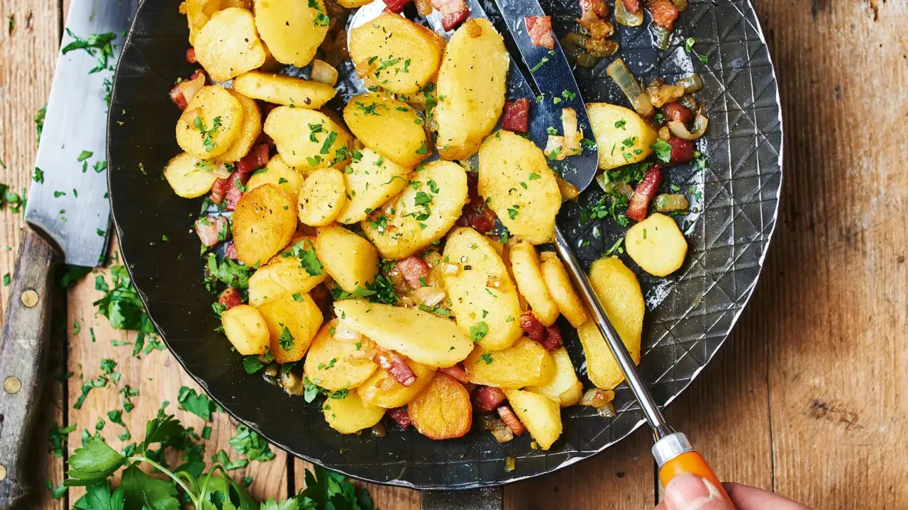 Bratkartoffeln mit mehligen Kartoffeln, Speck und Zwiebeln in einer Pfanne, garniert mit Petersilie.