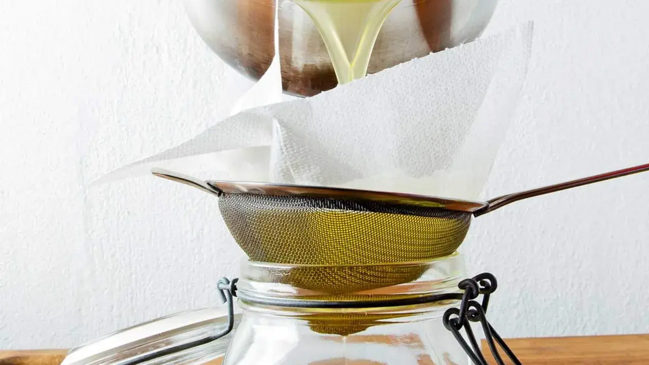 Flüssiges Ghee wird durch ein Sieb mit Küchenpapier in ein Glas gefiltert. So gelingt das Ghee selber machen.