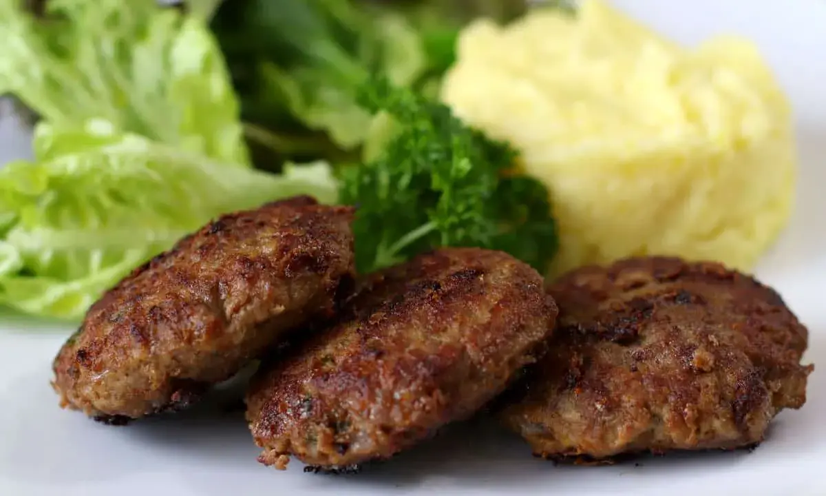 Drei goldbraune Fleischpflanzerl liegen auf einem Teller neben Kartoffelbrei und Salat. Ein leckeres Fleischpflanzerl Rezept zum Nachkochen.