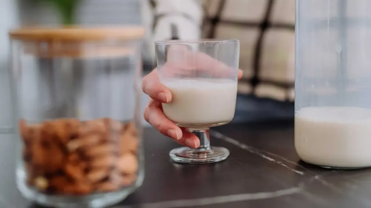 Glas Mandelmilch in Hand. Daneben ein Glas mit Mandeln und eine Flasche Milch. Man fragt sich, kann Mandelmilch schlecht werden?
