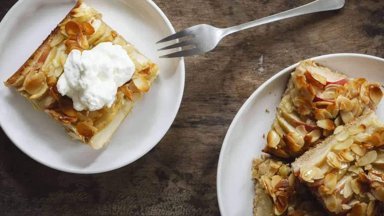 Zwei Teller mit saftigem Apfelkuchen vom Blech, bestreut mit Mandelsplittern. Eines der Stücke ist mit einer Sahnehaube garniert.
