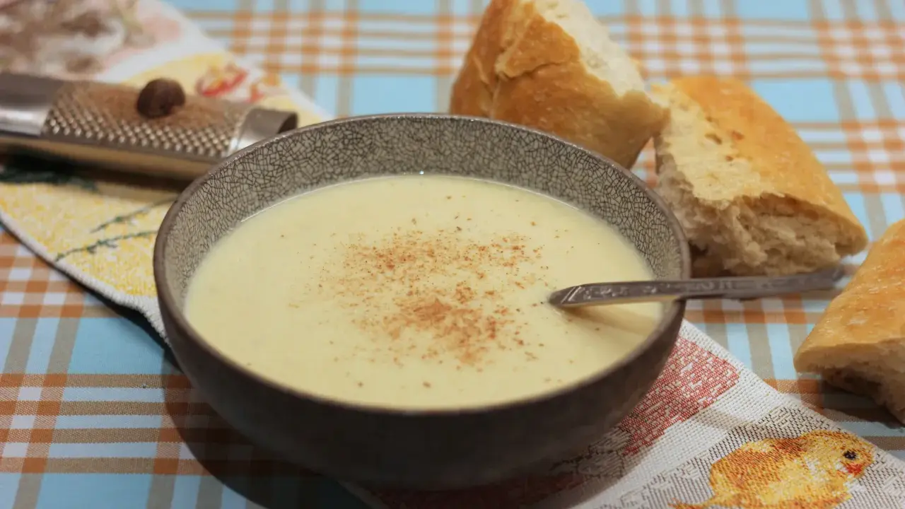 Eine Schüssel cremiger Stracciatella alla Romana, bestreut mit Zimt, neben knusprigem Brot. Ein Nussreibstein liegt auf einem bunten Tischtuch.
