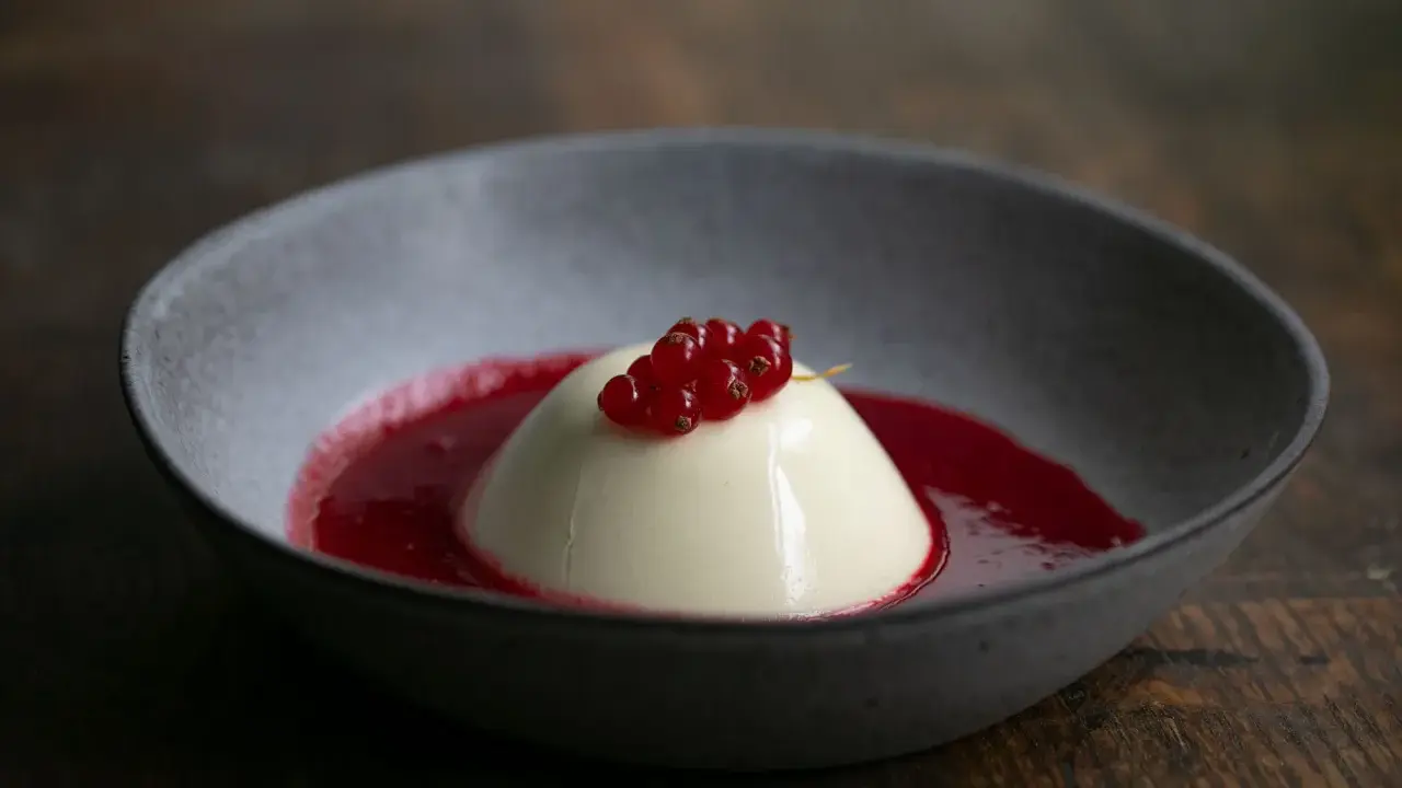 Cremige Panna Cotta mit roter Beerensauce und frischen Johannisbeeren in einer grauen Schale auf Holzhintergrund.
