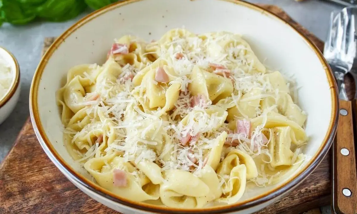 Eine Schüssel mit cremigen Tortellini alla panna, garniert mit geriebenem Parmesan und kleinen Schinkenwürfeln.