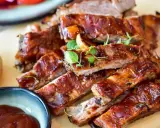 Saftige spareribs backofen, glasiert und mit Kräutern garniert, daneben eine Schale BBQ-Sauce und eine Kartoffel.