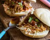 Saftiges pulled pork auf einem Brötchen mit Coleslaw und BBQ-Sauce, garniert mit Frühlingszwiebeln. Ein Löffel mit Sauce liegt daneben.