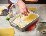 Hände legen Lasagneplatten auf eine Schicht Käse und Sauce. Ein Schritt im Lasagne rezept.