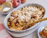 Ein goldbrauner Pfirsich Crumble mit knuspriger Streuseldecke, bestäubt mit Puderzucker. Ein Löffel steckt darin, bereit zum Genießen.