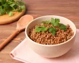 Schüssel mit Buchweizen als Beilage, garniert mit Petersilie. Daneben ein Holzlöffel und ein Bund frischer Kräuter.