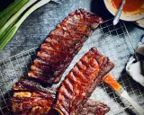 Saftige spare ribs, glasiert mit BBQ-Sauce, auf einem Grillrost. Daneben Frühlingszwiebeln und eine Schüssel Sauce.