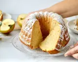 Ein saftiger Kuchen mit Apfelmus, bestäubt mit Puderzucker. Daneben liegen frische Apfelhälften und ein Glas Apfelmus.