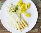 Ein Teller mit weißem Spargel, garniert mit Petersilie und überzogen mit cremiger Hollandaise. Dazu gibt es kleine, goldgelbe Kartoffeln.