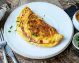 Goldbraunes Omelett auf weißem Teller, garniert mit Schnittlauch. Daneben Schüssel mit geriebenem Käse und gehacktem Schnittlauch. Perfekt für ein schnelles Omelette Rezept.