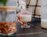Glas Mandelmilch in Hand. Daneben ein Glas mit Mandeln und eine Flasche Milch. Man fragt sich, kann Mandelmilch schlecht werden?