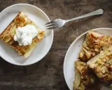 Zwei Teller mit saftigem Apfelkuchen vom Blech, bestreut mit Mandelsplittern. Eines der Stücke ist mit einer Sahnehaube garniert.