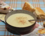 Eine Schüssel cremiger Stracciatella alla Romana, bestreut mit Zimt, neben knusprigem Brot. Ein Nussreibstein liegt auf einem bunten Tischtuch.