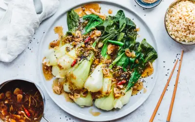 Ein Teller mit gebratenem Pak Choi, garniert mit Sesam und Chili, serviert mit Reis und einer Sauce.