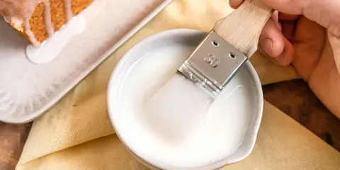 Ein Pinsel taucht in eine Schüssel mit zuckerguss, bereit, einen warmen oder kalten Kuchen zu verfeinern.