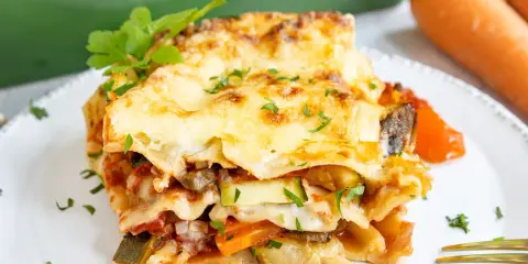 Ein Stück herzhafte vegetarische Lasagne mit geschmolzenem Käse und frischer Petersilie, daneben Karotten.