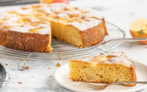 Ein saftiger glutenfreier Apfelkuchen, bestäubt mit Puderzucker, auf einem Gitterrost. Ein Stück auf einem Teller mit Gabel daneben.