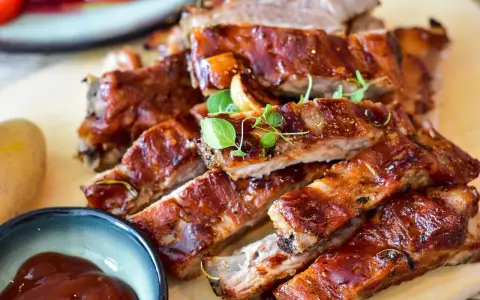 Saftige spareribs backofen, glasiert und mit Kräutern garniert, daneben eine Schale BBQ-Sauce und eine Kartoffel.