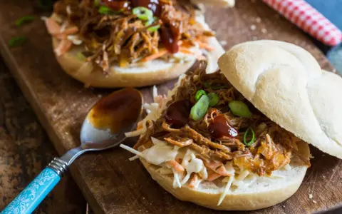 Saftiges pulled pork auf einem Brötchen mit Coleslaw und BBQ-Sauce, garniert mit Frühlingszwiebeln. Ein Löffel mit Sauce liegt daneben.