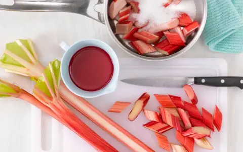 Frischer Rhabarber wird geschnitten und mit Zucker für ein leckeres Rhabarberkompott vorbereitet. Ein Messer und eine Schüssel mit rotem Saft liegen bereit.