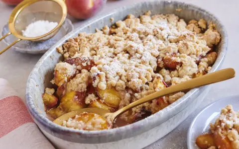 Ein goldbrauner Pfirsich Crumble mit knuspriger Streuseldecke, bestäubt mit Puderzucker. Ein Löffel steckt darin, bereit zum Genießen.