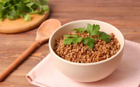 Schüssel mit Buchweizen als Beilage, garniert mit Petersilie. Daneben ein Holzlöffel und ein Bund frischer Kräuter.