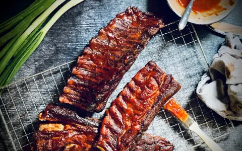 Saftige spare ribs, glasiert mit BBQ-Sauce, auf einem Grillrost. Daneben Frühlingszwiebeln und eine Schüssel Sauce.