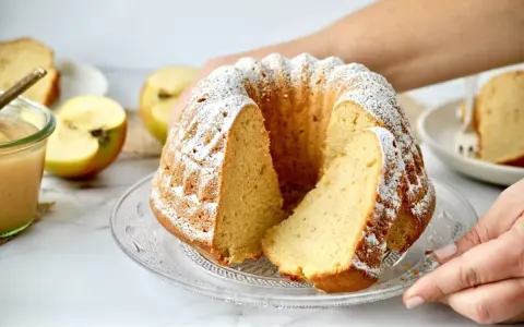 Ein saftiger Kuchen mit Apfelmus, bestäubt mit Puderzucker. Daneben liegen frische Apfelhälften und ein Glas Apfelmus.