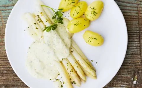 Ein Teller mit weißem Spargel, garniert mit Petersilie und überzogen mit cremiger Hollandaise. Dazu gibt es kleine, goldgelbe Kartoffeln.