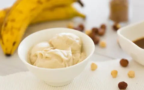 Cremiges Bananeneis, gemacht aus gefrorenen Bananen, in einer Schüssel. Daneben liegen Nüsse und ein Schälchen mit Schokoladensauce.