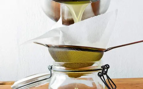 Flüssiges Ghee wird durch ein Sieb mit Küchenpapier in ein Glas gefiltert. So gelingt das Ghee selber machen.