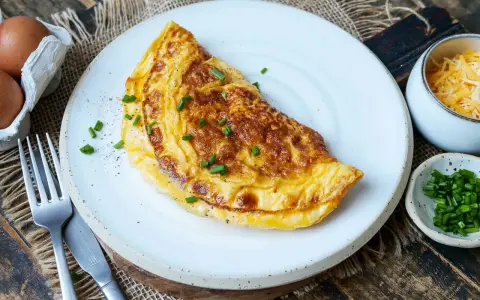 Goldbraunes Omelett auf weißem Teller, garniert mit Schnittlauch. Daneben Schüssel mit geriebenem Käse und gehacktem Schnittlauch. Perfekt für ein schnelles Omelette Rezept.