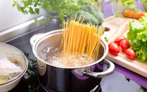 Spaghetti kochen: Lange Nudeln werden in kochendes Wasser gegeben. Frisches Gemüse wie Brokkoli, Karotten und Tomaten liegen bereit.