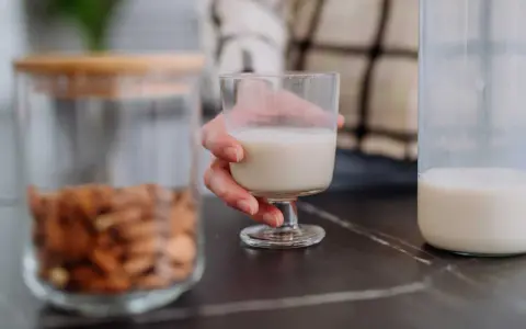 Glas Mandelmilch in Hand. Daneben ein Glas mit Mandeln und eine Flasche Milch. Man fragt sich, kann Mandelmilch schlecht werden?