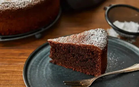Ein Stück saftiger Schokoladenkuchen ohne Mehl, bestäubt mit Puderzucker, liegt auf einem dunklen Teller. Daneben eine Gabel und ein Sieb mit Puderzucker.